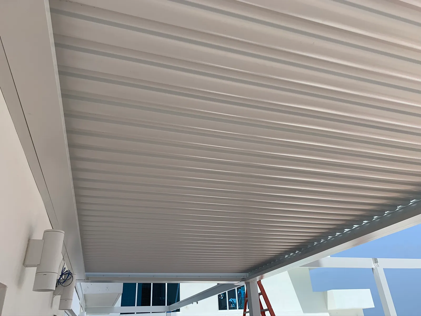 Underside of a white louvered roof with louvers closed, wall lights on the left.
