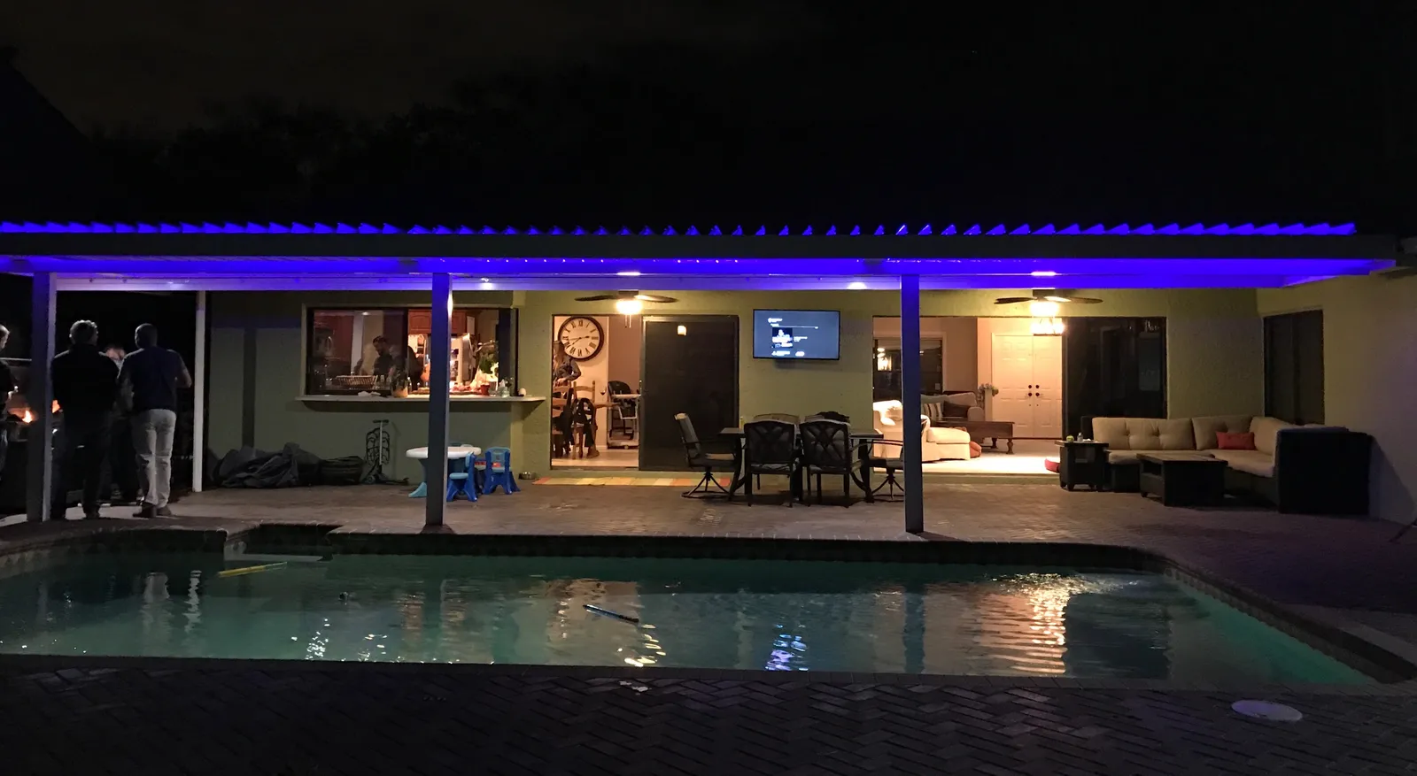 Long louvered roof edged with blue LED lighting over a patio beside a pool at night