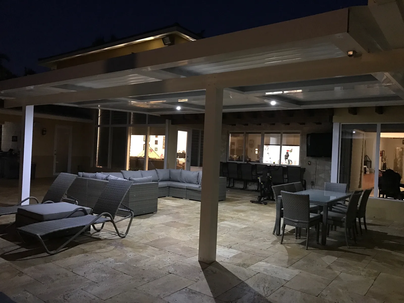 Louvered roof with white recessed lights over a travertine patio with lounge and dining furniture at night