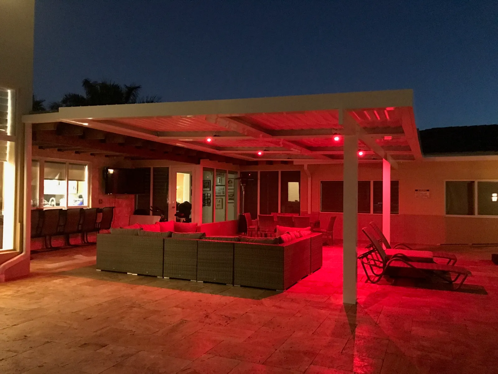White louvered roof lit with red LED lights over a travertine patio with wicker seating at dusk