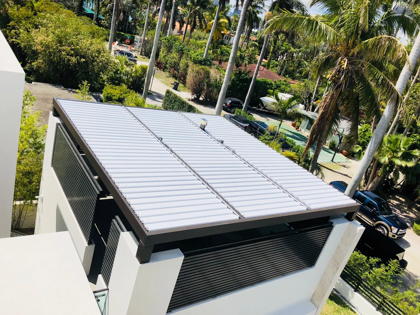 Closed white louvered roof with bronze frame on a rooftop terrace seen from above among palm trees