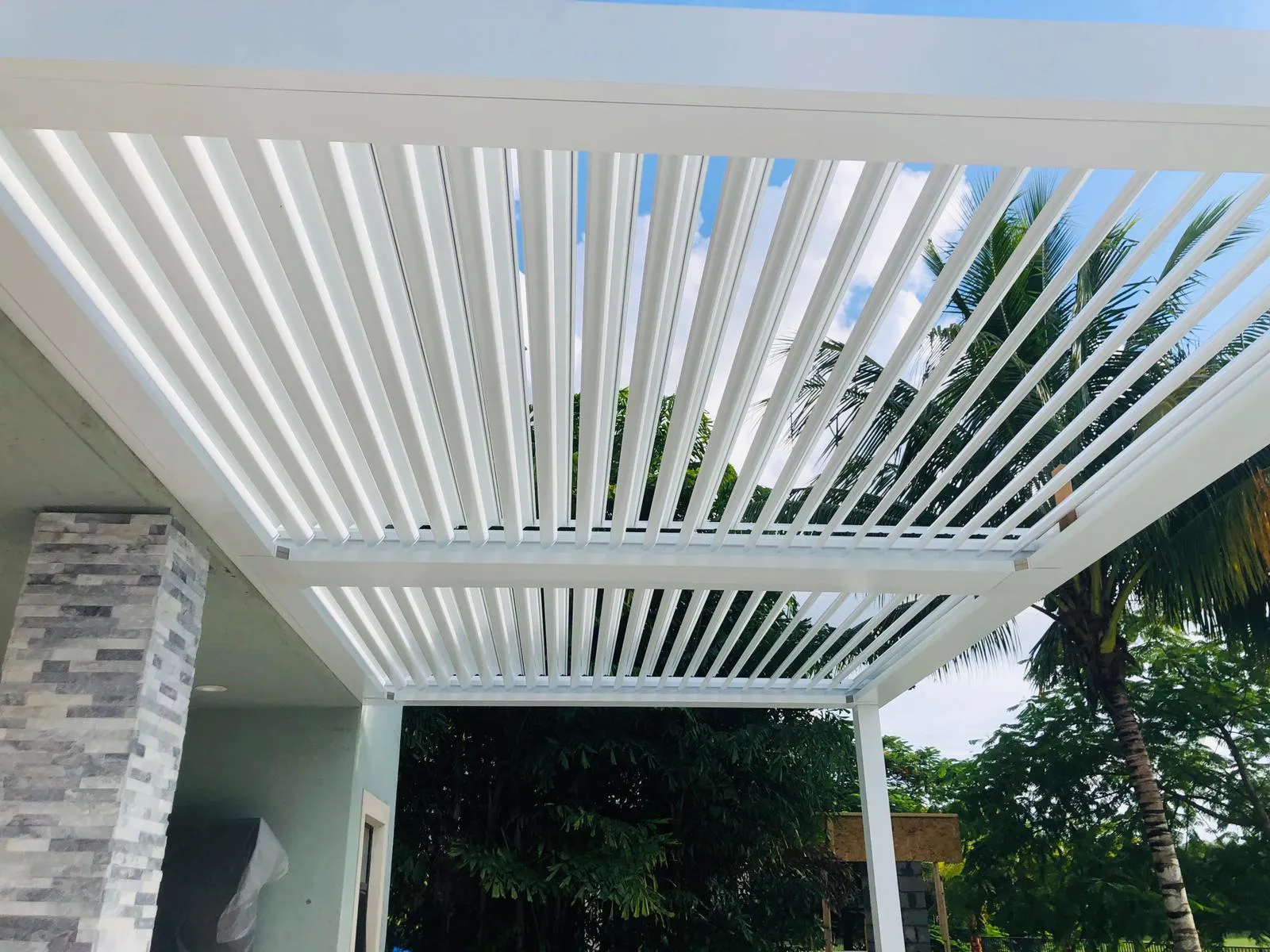 White louvered roof blades tilted open seen from below with palm trees and sky beyond