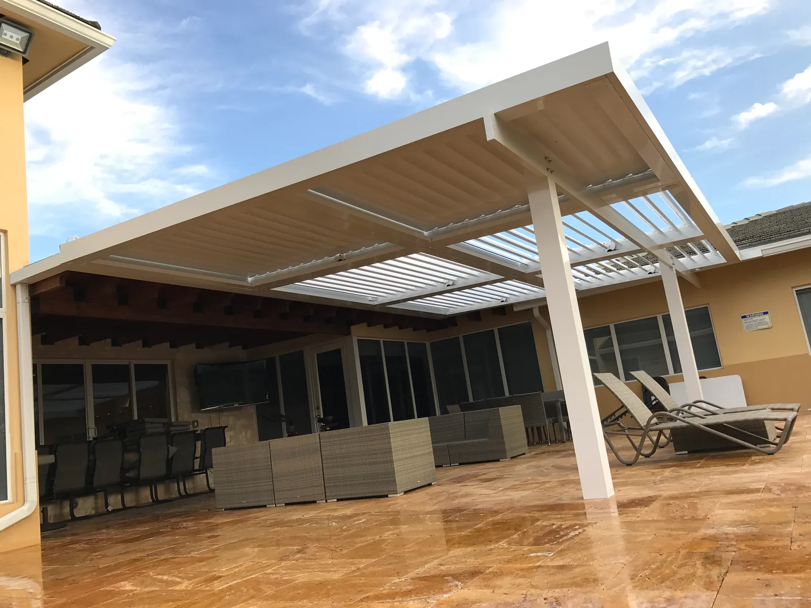Large white louvered roof with partly open blades over a travertine patio with wicker lounge furniture