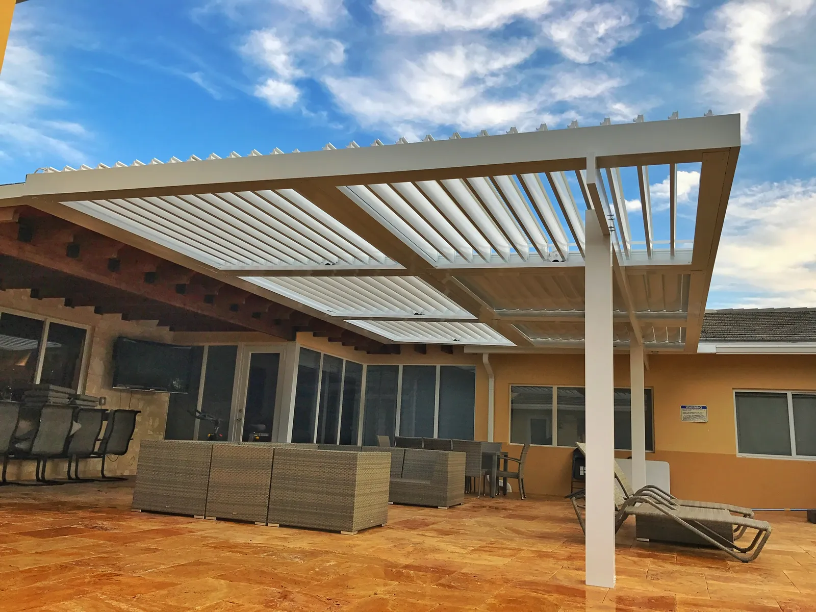 White louvered roof with blades open against a blue sky over a travertine patio with wicker furniture