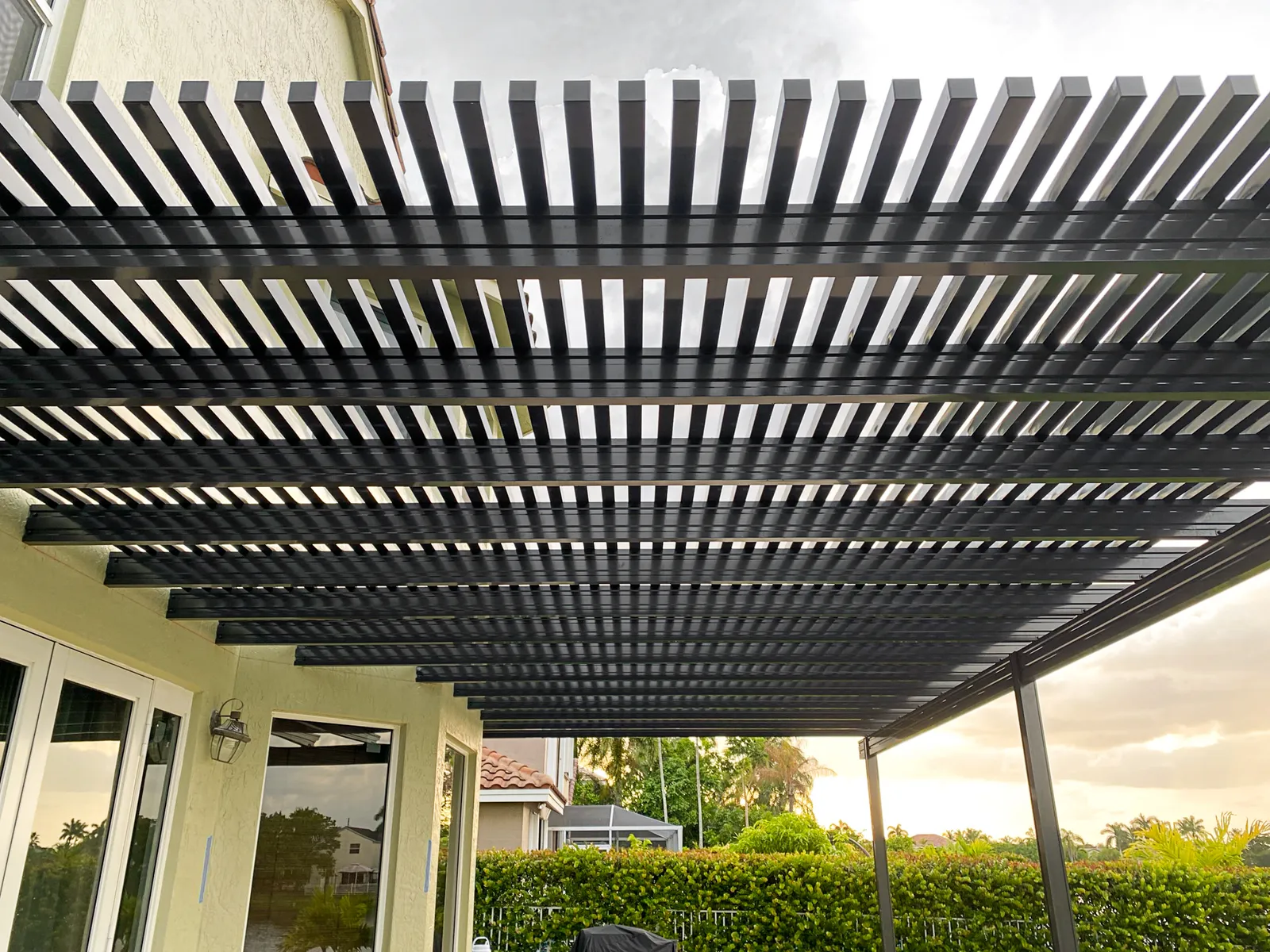 Underside of a black aluminum lattice pergola attached to a house with a sunset sky behind