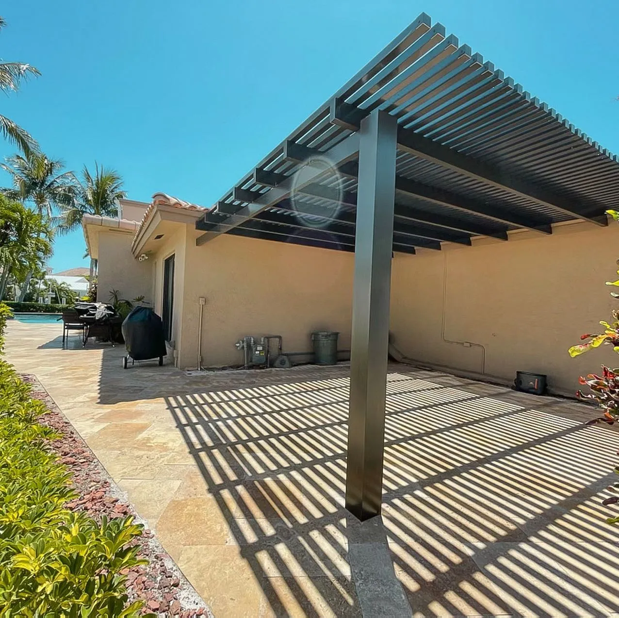 Dark bronze aluminum lattice pergola on a single corner post casting striped shadows on a travertine patio