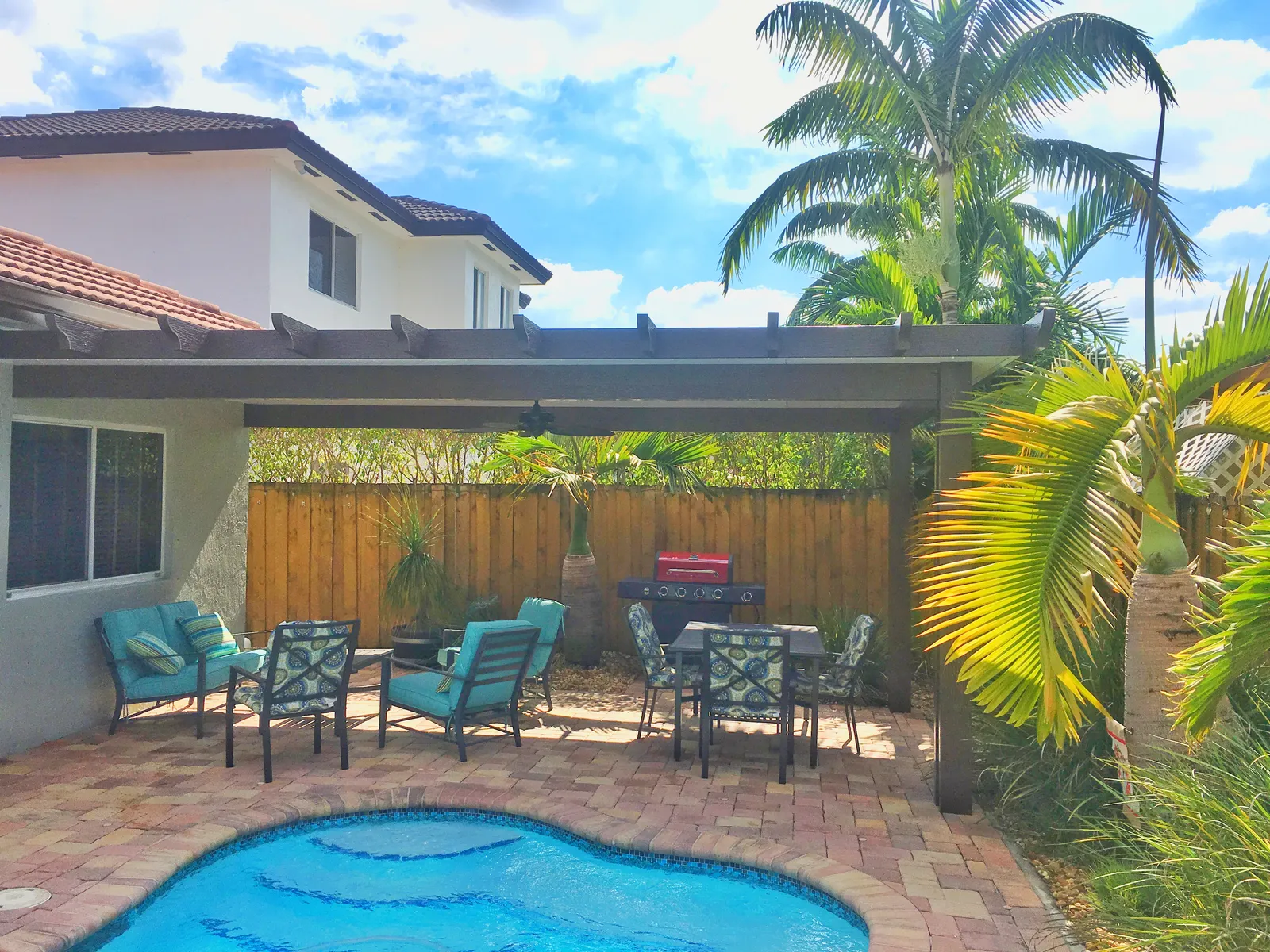 Brown covered pergola with rafter tails over a paver patio with turquoise seating, grill and freeform pool