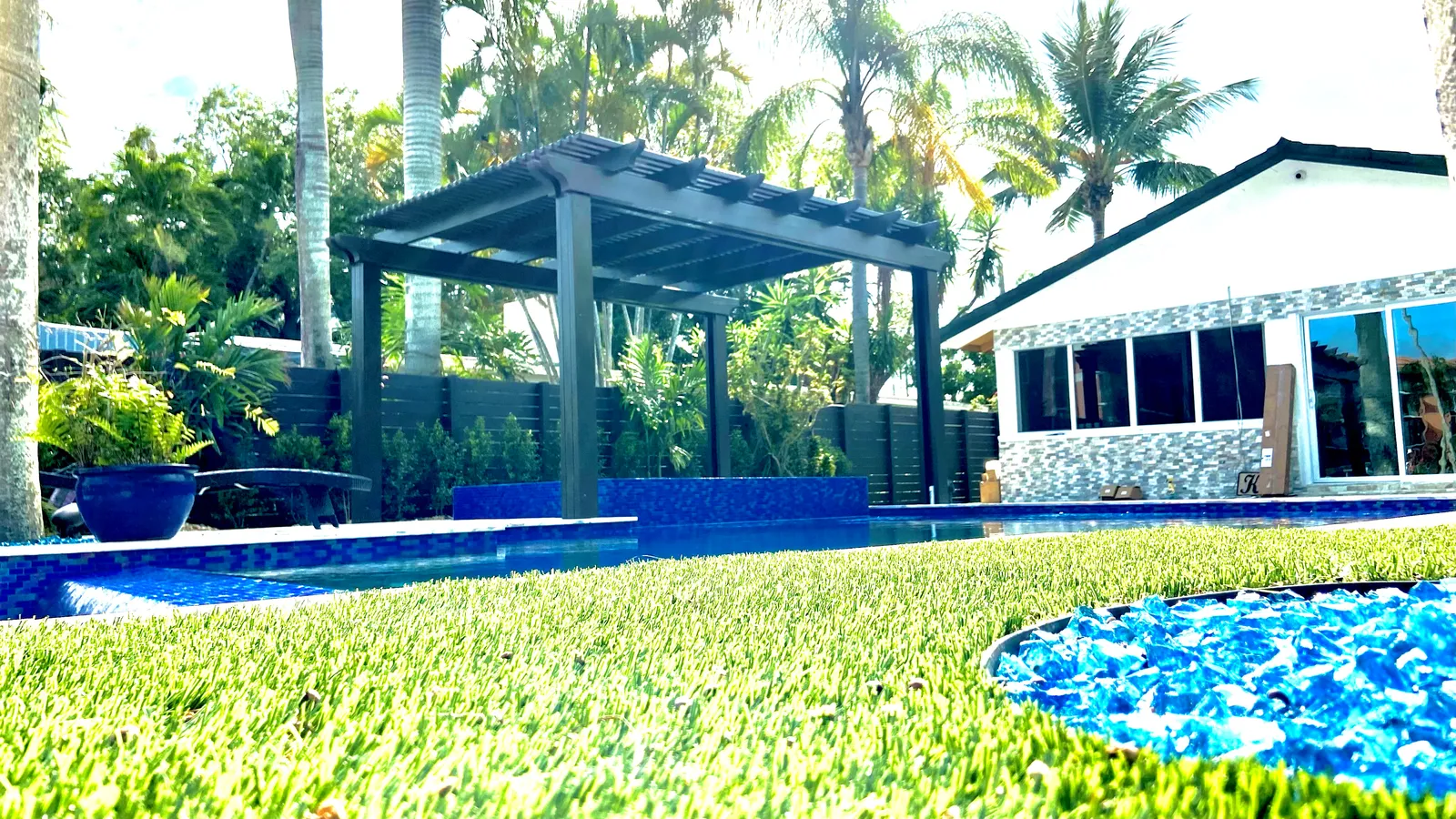 Low view across artificial turf to a dark bronze pergola over a spa with a white house behind