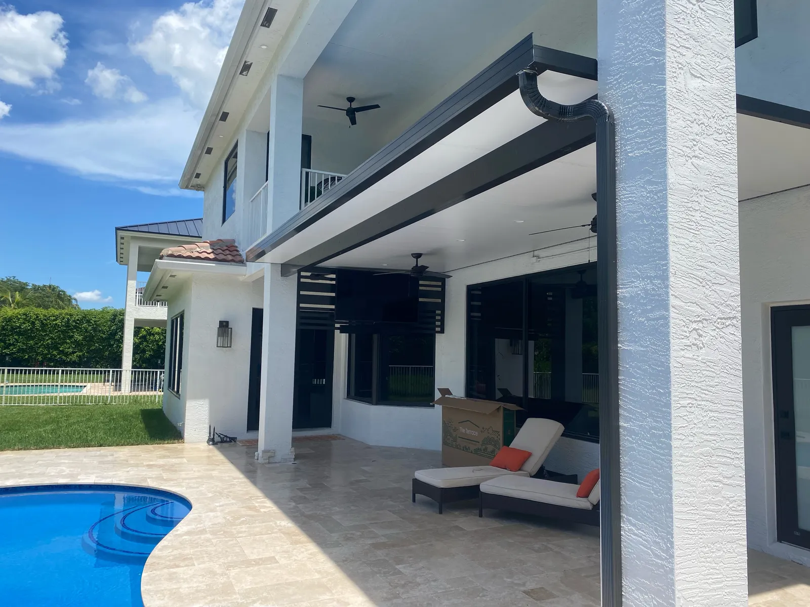 Finished black-fascia insulated patio cover with ceiling fans on a white two-story home beside a pool