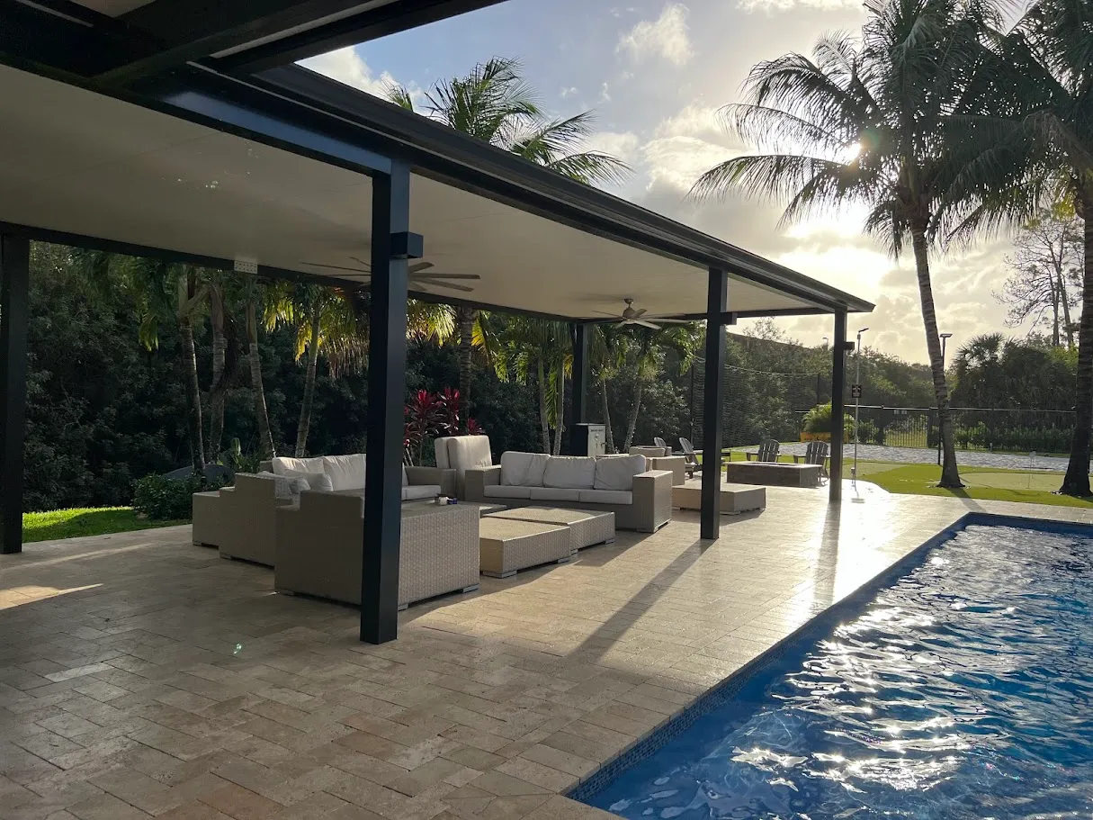 Black-framed insulated patio cover with ceiling fans and wicker sofas beside a lap pool at sunset