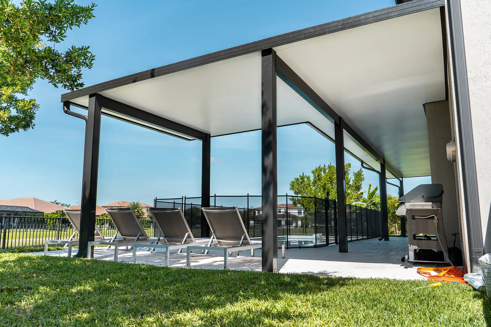 Black-framed insulated patio cover over four sun loungers seen from the lawn under a clear blue sky