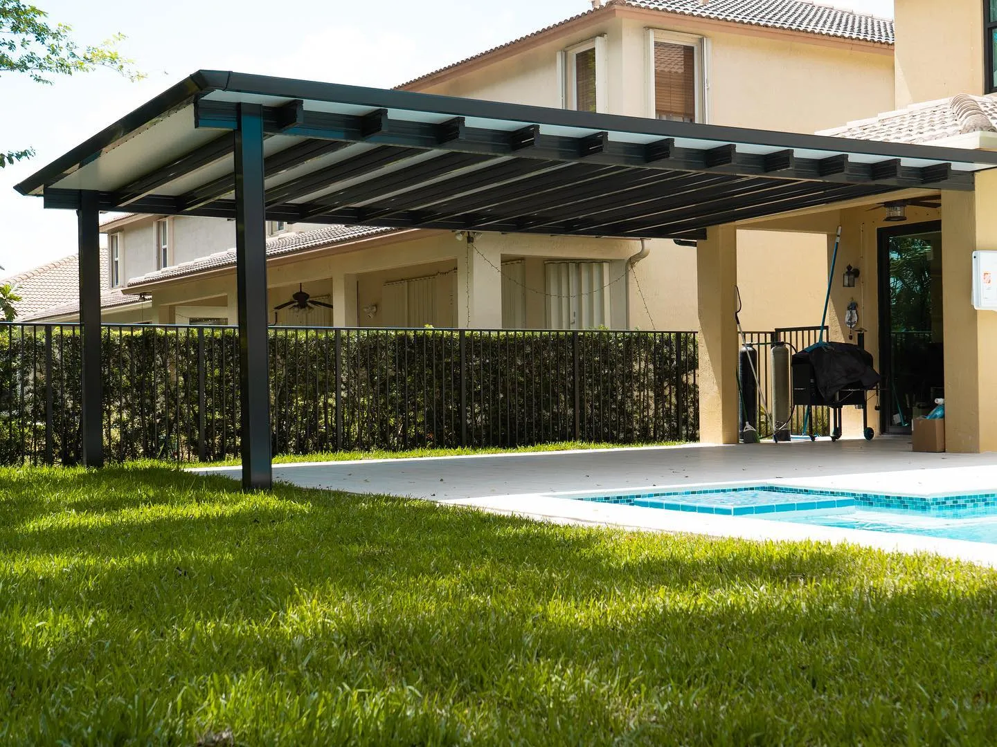 Black-framed insulated patio cover with rafter tails extending from a yellow two-story home beside a pool