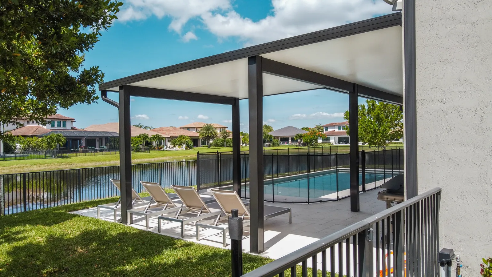 Low aerial view of a black-framed insulated patio cover over loungers beside a fenced pool and lake