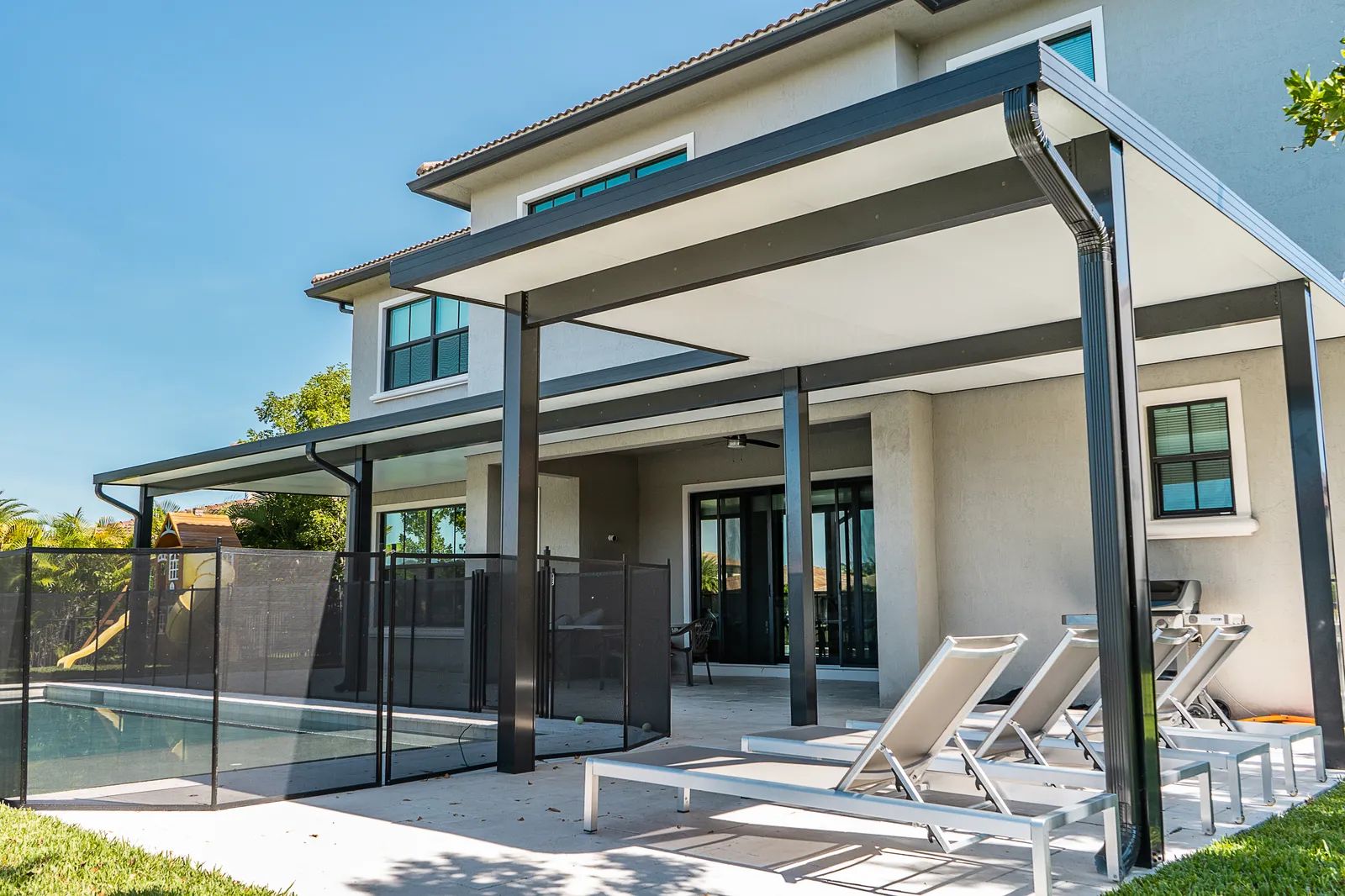 Black-framed insulated patio cover with downspout over sun loungers on a two-story home by the pool