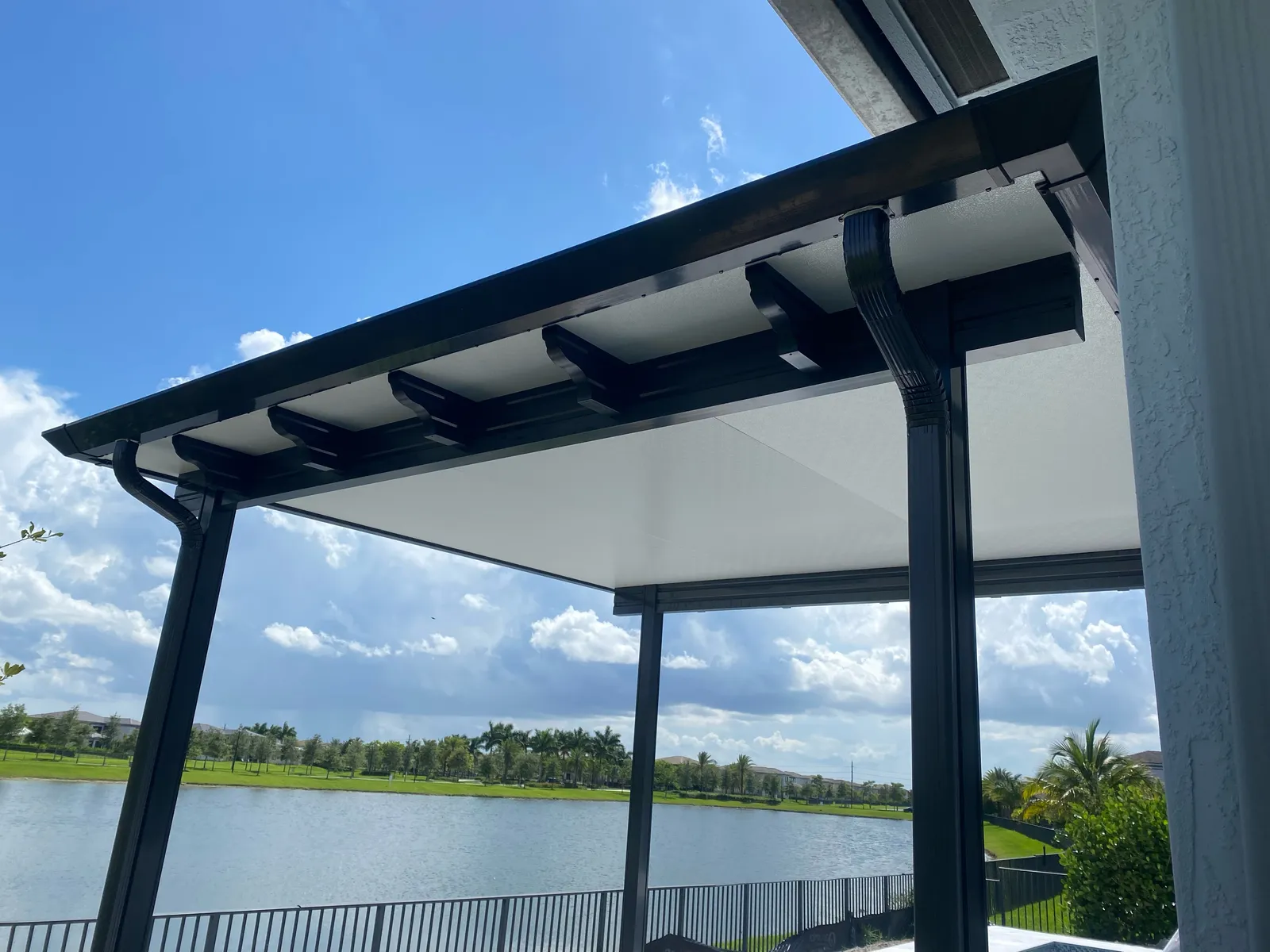 Upward view of a black patio cover with scalloped rafter tails, gutter and downspout framing a lake
