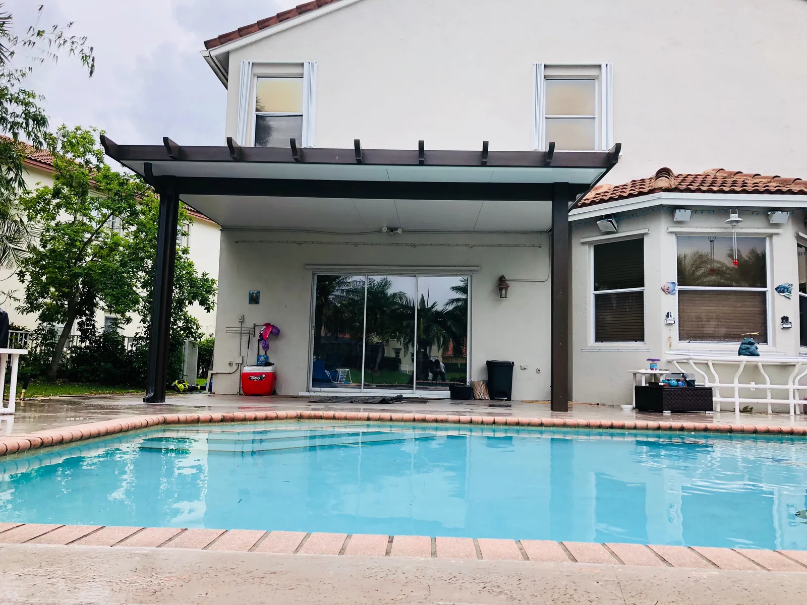 Brown insulated patio cover with rafter tails on a two-story home seen across a pool with brick coping
