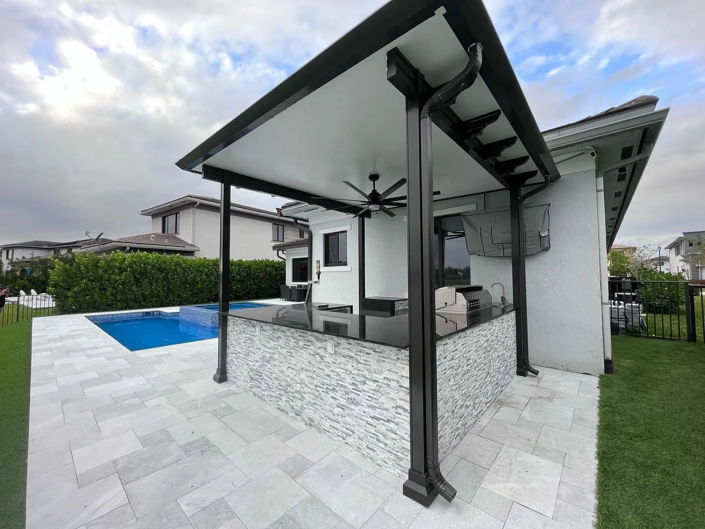 Black aluminum insulated patio cover over a stacked stone outdoor kitchen next to a pool.