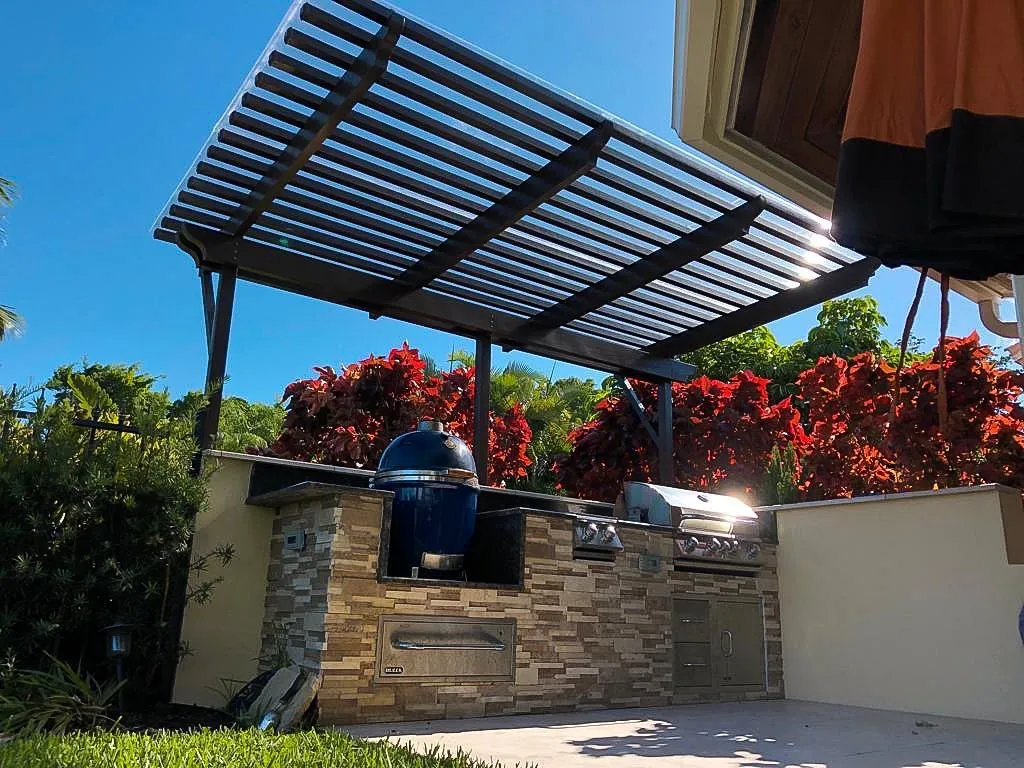 Dark lattice pergola over a stacked-stone outdoor kitchen with kamado grill and gas grill, red crotons behind