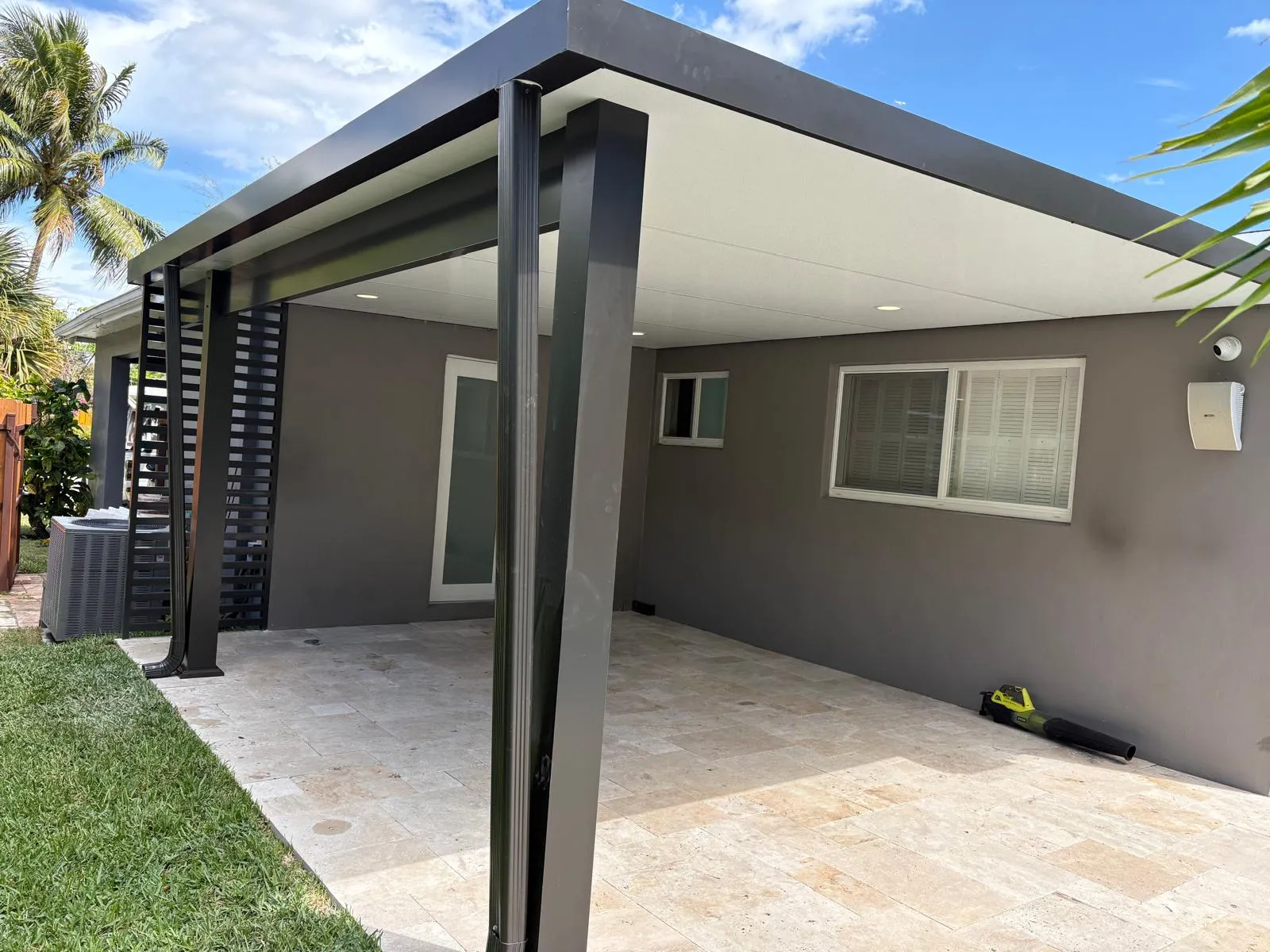 Black-fascia flat insulated patio cover with slatted privacy panel on a gray stucco house over a travertine patio