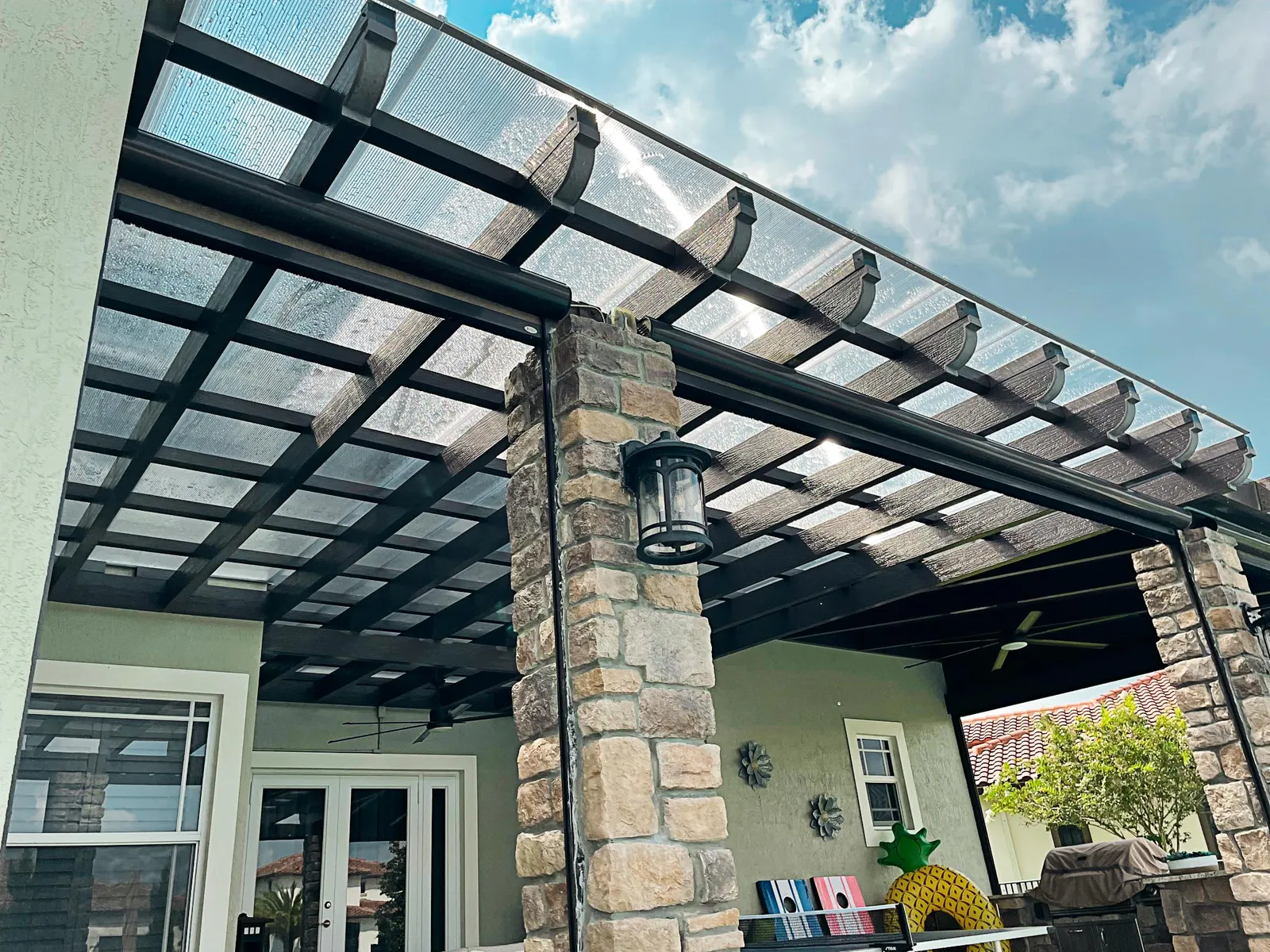 Dark woodgrain pergola with clear polycarbonate roof and scalloped rafter tails on stacked-stone columns