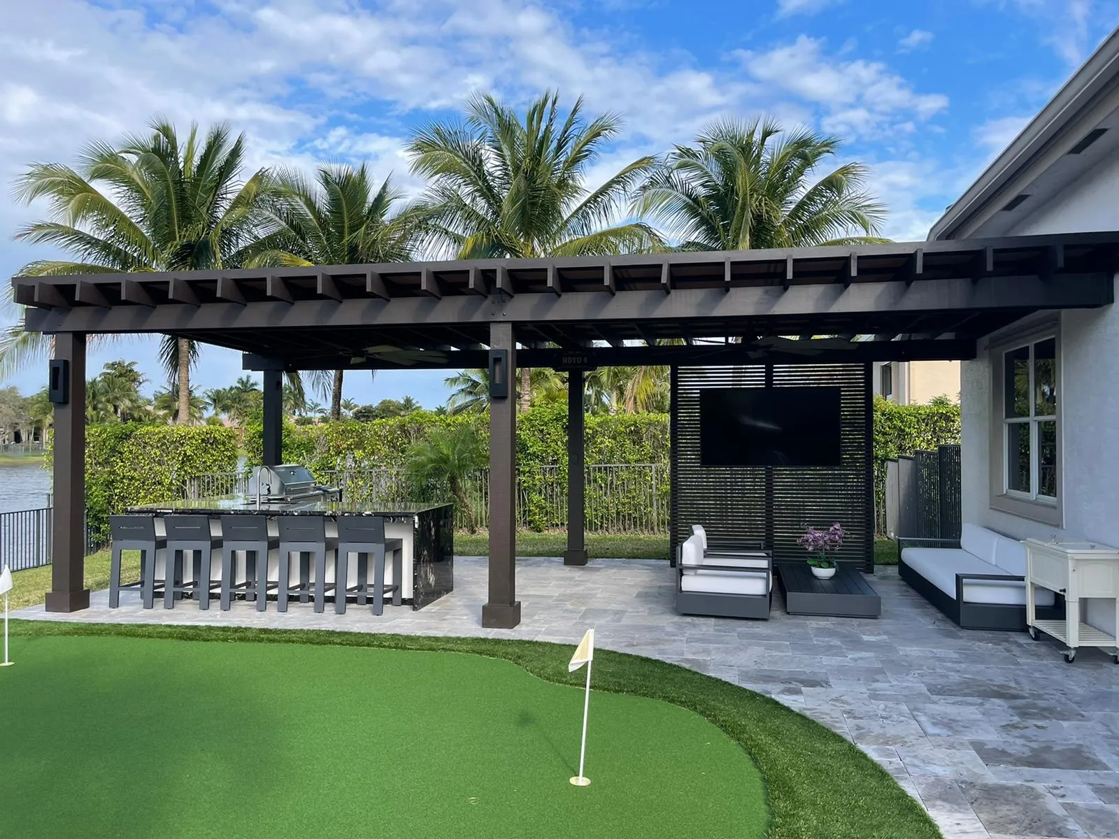 Dark wood pergola over an outdoor kitchen bar and lounge with TV screen beside a putting green.