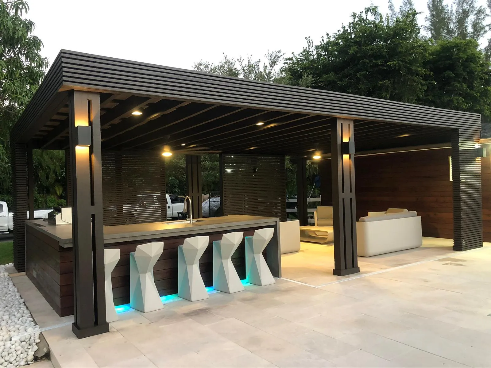 Dark wood pavilion with outdoor bar, sculptural stools, LED toe-kick lights and lounge at dusk.