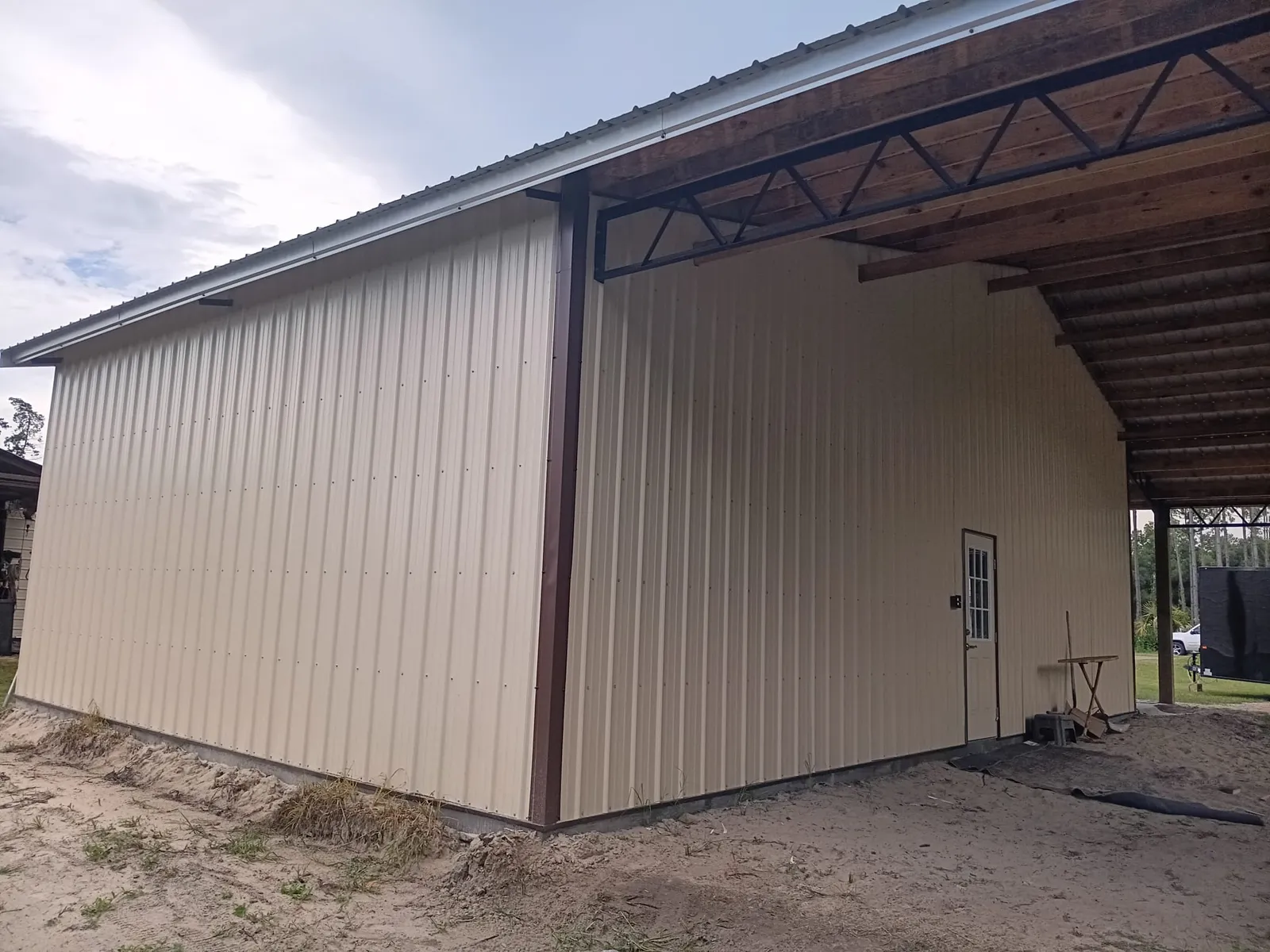 Enclosed pole barn with tan metal siding and brown trim next to an open lean-to with steel trusses.