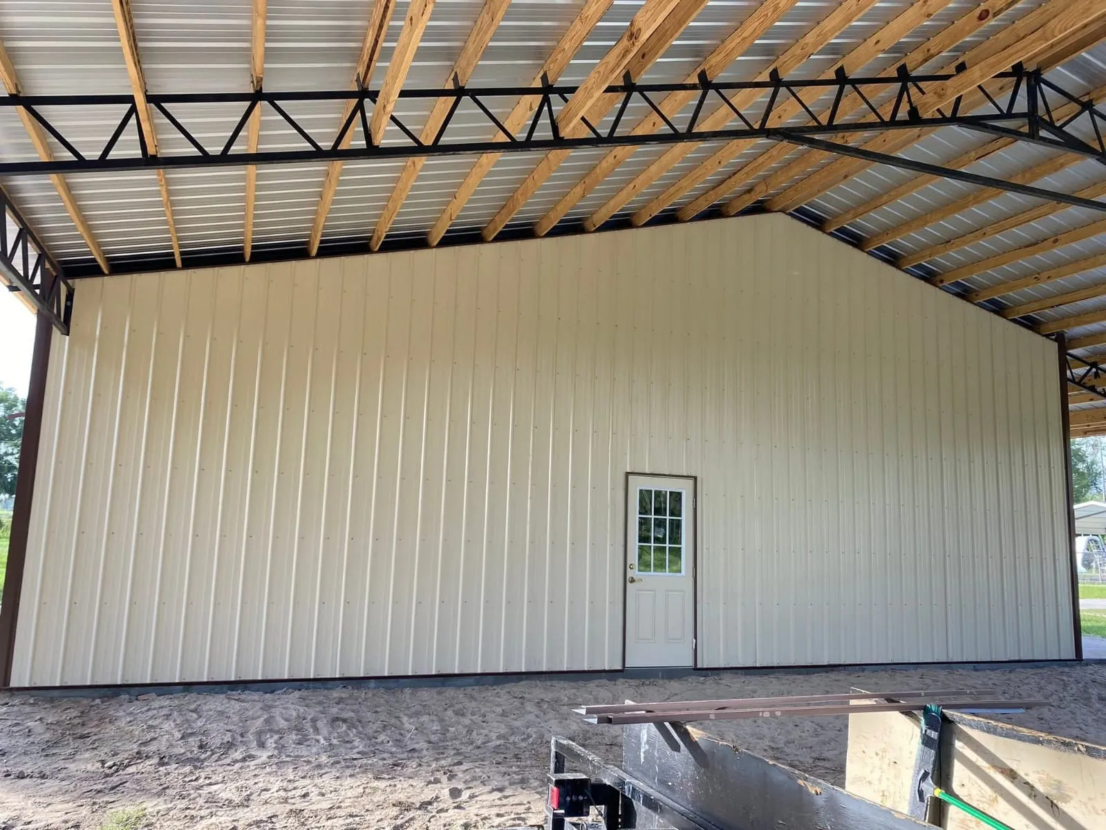 Tan metal gable wall with a walk door under a pole barn lean-to roof with black steel trusses.