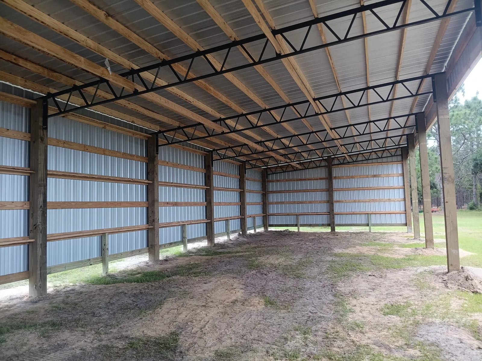 Long three-sided pole barn with wood posts, black steel trusses and metal walls on a dirt floor.