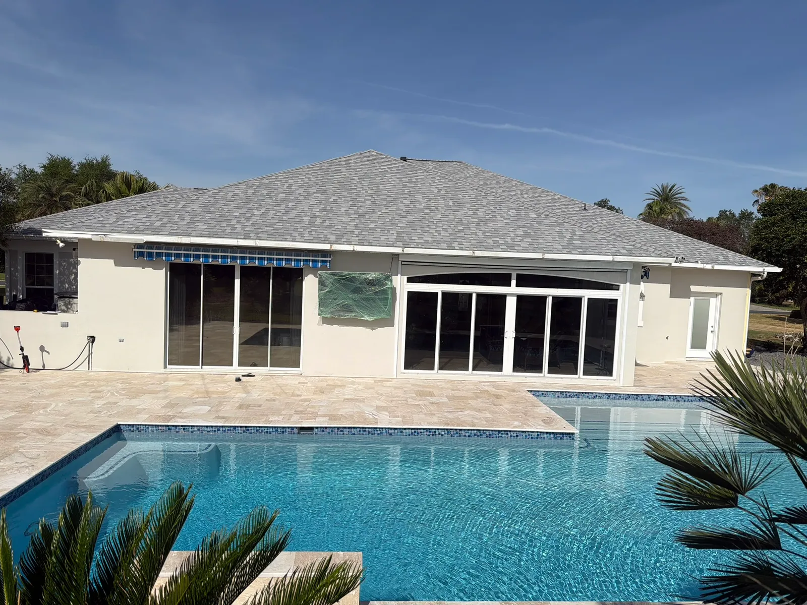 Remodeled pool with blue glass waterline tile and ivory travertine deck behind a stucco house.