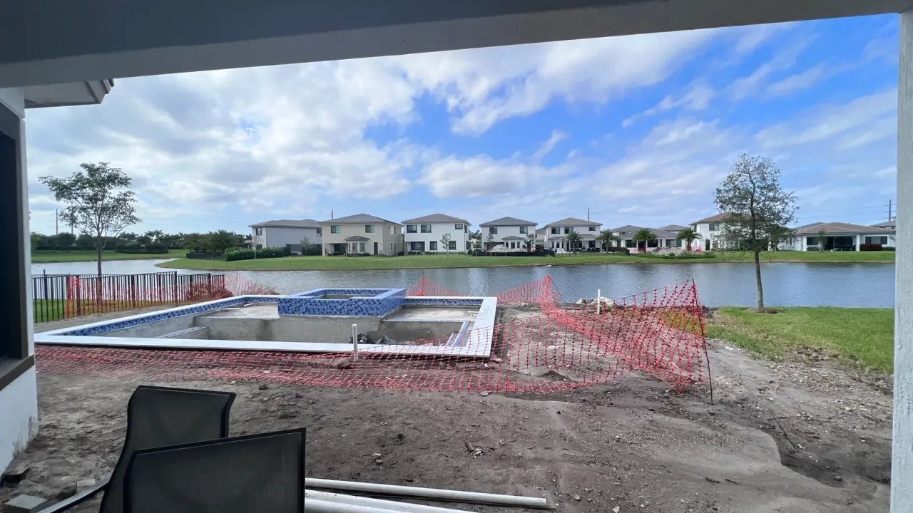 Pool under construction on a lakefront lot, with a blue glass tile spa and orange safety fencing, seen from a covered porch.