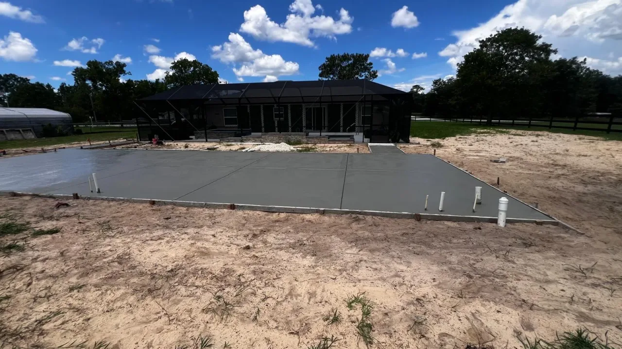Freshly poured concrete slab with edge forms and plumbing stubs, in front of a black screen enclosure on a sandy lot.