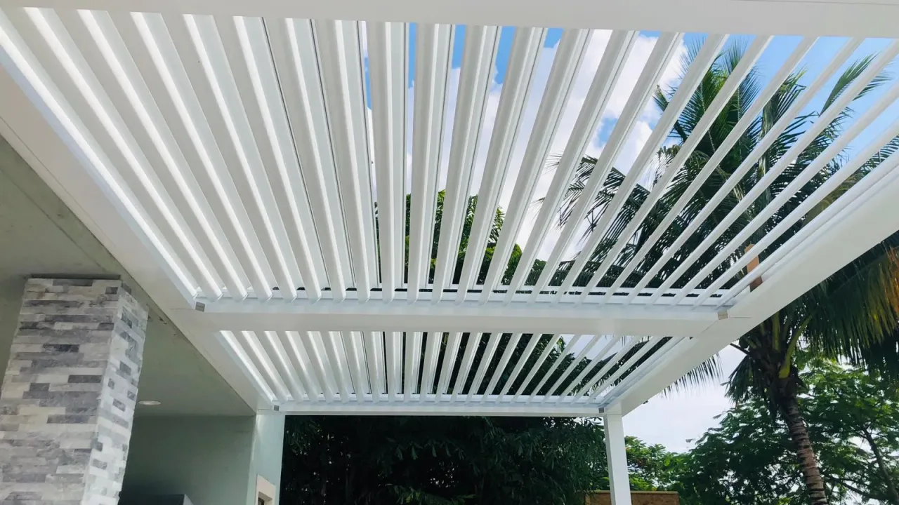 White louvered roof blades tilted open, seen from below against the sky beside a stacked-stone column and palms