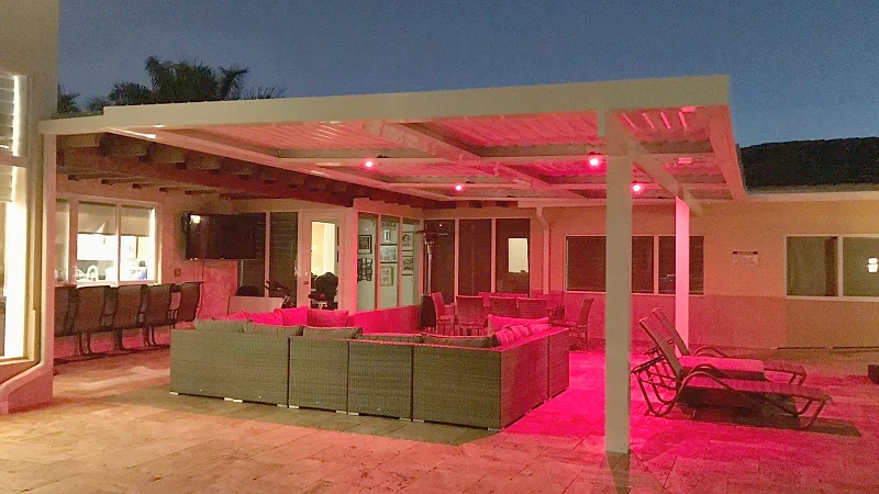 White louvered roof lit with red LEDs at dusk over a wicker sectional and lounge chairs on a travertine patio