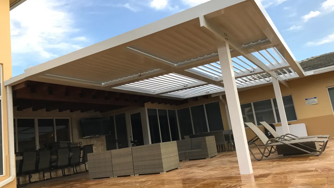 White louvered roof with blades partly open over wicker seating and loungers on wet stone paving, a TV on the wall behind