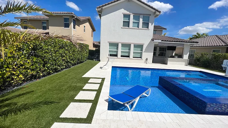 Pool with a raised dark-tile spa, a floating lounger and stepping pads set in artificial turf behind a two-story house