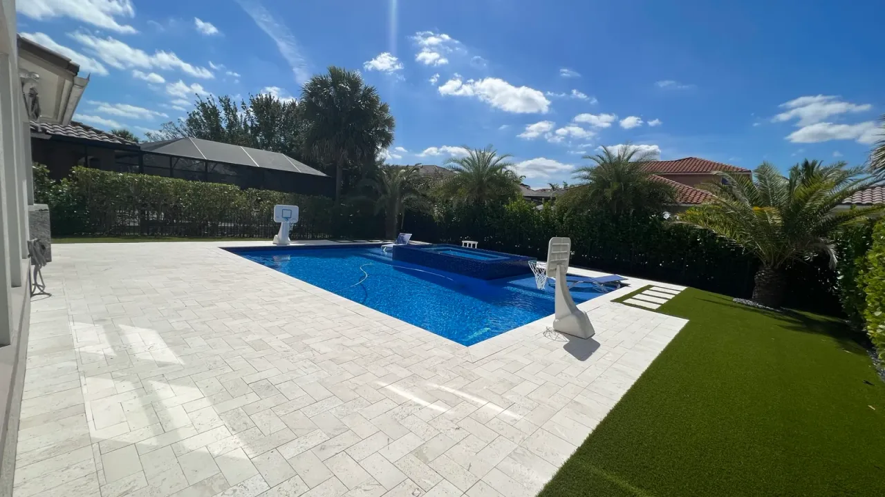 Pool with deep blue water and a blue-tiled raised spa, framed by a wide ivory travertine deck and a strip of artificial turf.