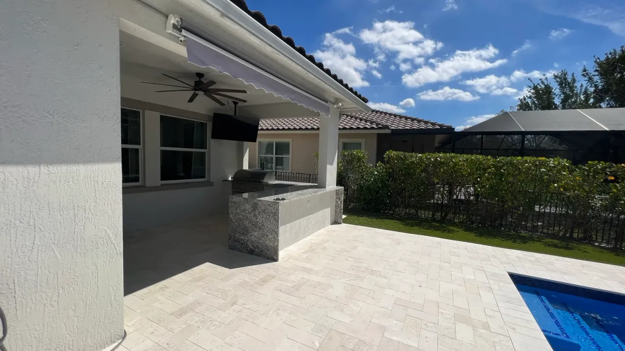 Outdoor kitchen island at the edge of a covered patio, with a ceiling fan and awning above and a pool corner in the sun.