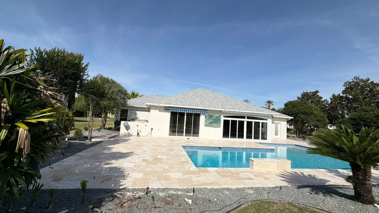 Finished pool with a raised water-feature wall at its edge, a wide travertine deck reaching the house and gray gravel beds.