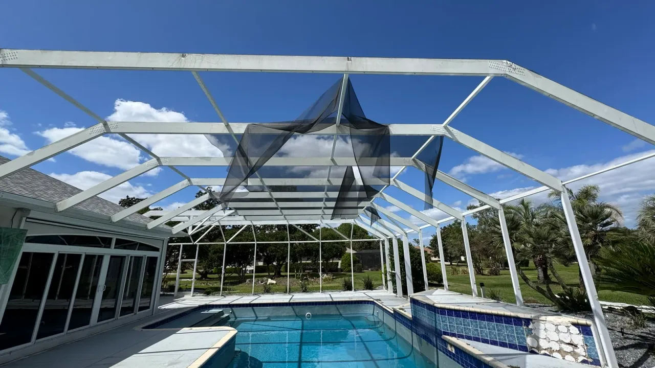 Screen enclosure with torn mesh hanging from its frame over a blue-tiled pool, with tiles missing from a raised wall.