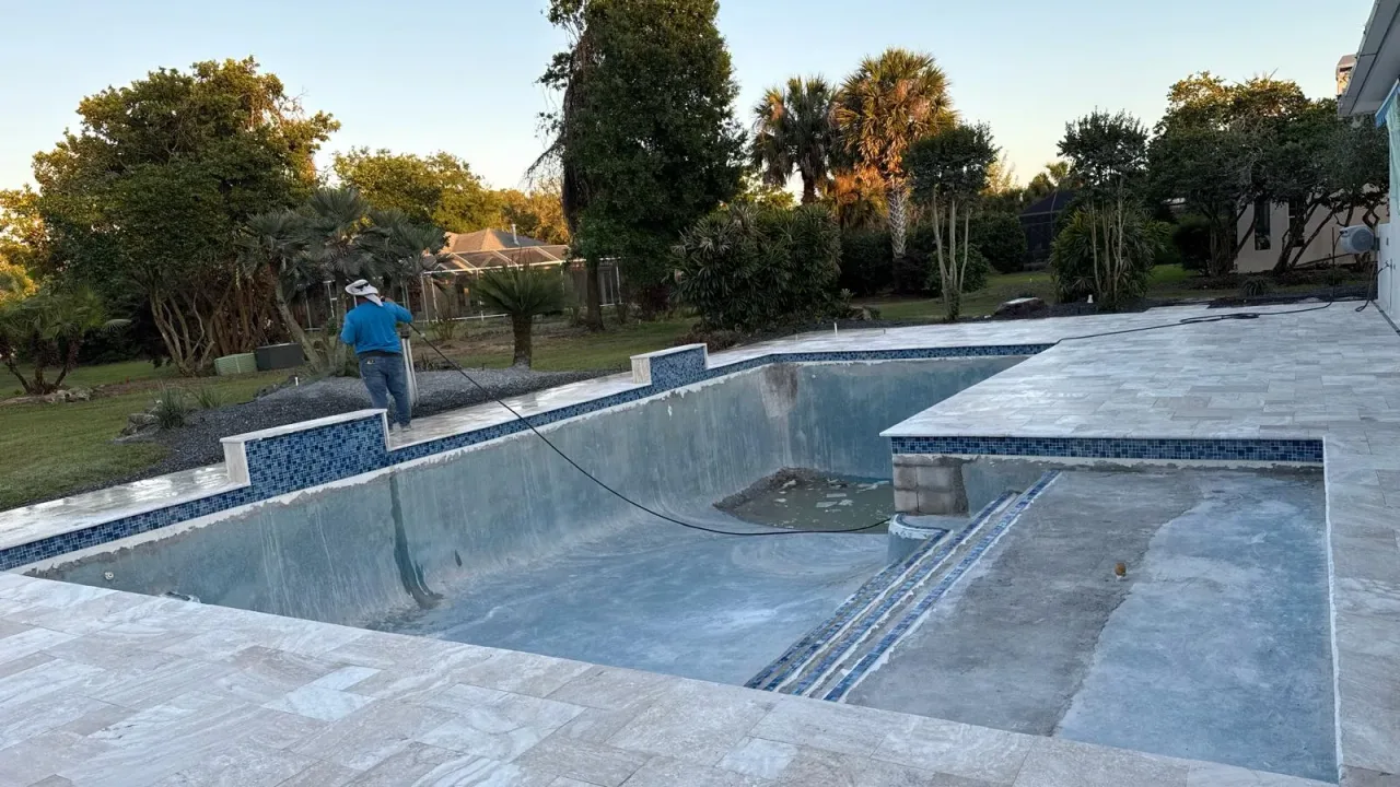 Empty pool with a wide, still-unfinished sun shelf at one end, new blue mosaic waterline tile and a travertine deck.