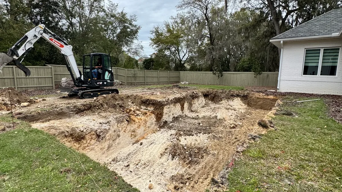 Mini excavator beside a freshly dug pool hole in a backyard enclosed by a wooden fence, with mature trees behind.