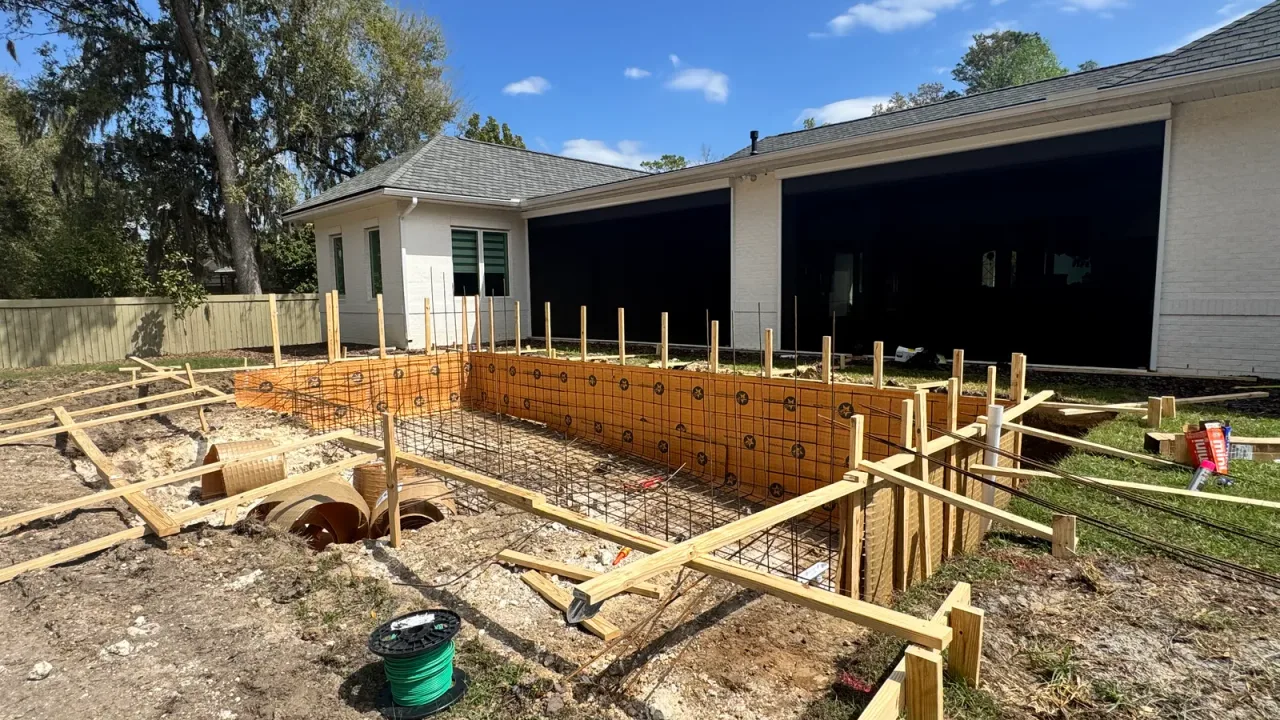 Steel rebar grid and wooden formwork for a new pool laid out close behind a white brick house.