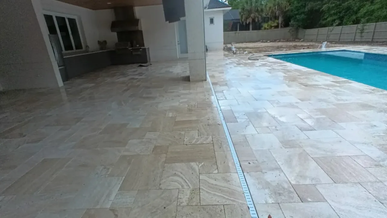 Travertine paving running from a covered patio to the pool deck, with a linear drain set along the joint.