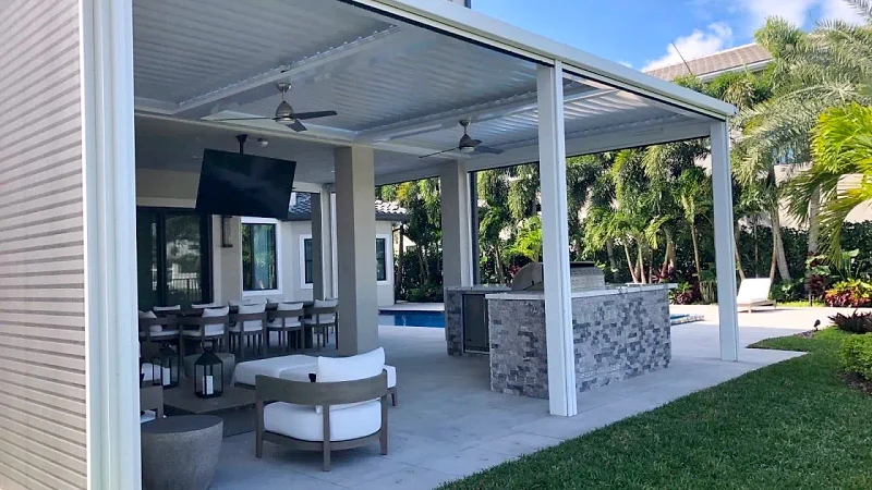 White louvered roof with ceiling fans and a TV over a stacked-stone kitchen island, lounge chairs and a dining table