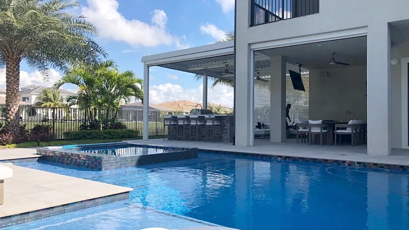 Raised spa in iridescent glass tile in front of a white louvered roof sheltering an outdoor bar and a covered dining area