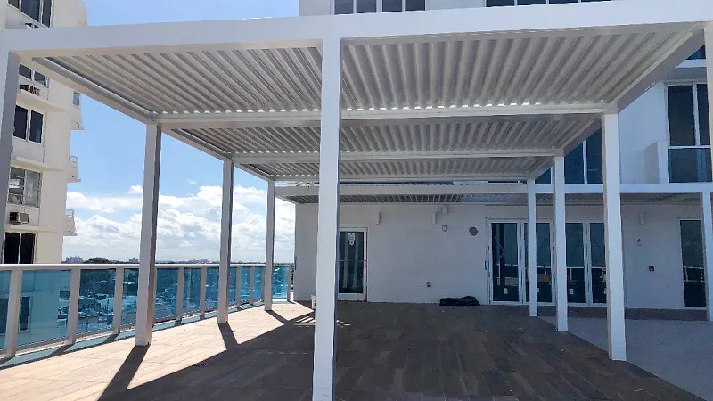 White louvered pergolas over a wood-look deck on a high-rise terrace with a glass railing, apartment towers on both sides