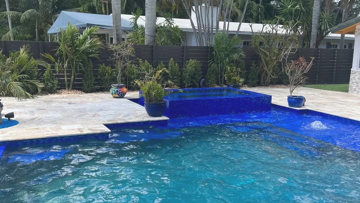 Raised spa clad in cobalt blue mosaic at the back of a deep blue pool, with a stone deck and potted plants.