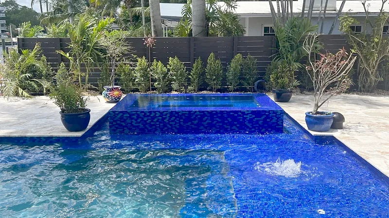 Cobalt glass mosaic pool with a bubbler and a raised mosaic spa, potted plants and palms behind a dark slatted fence