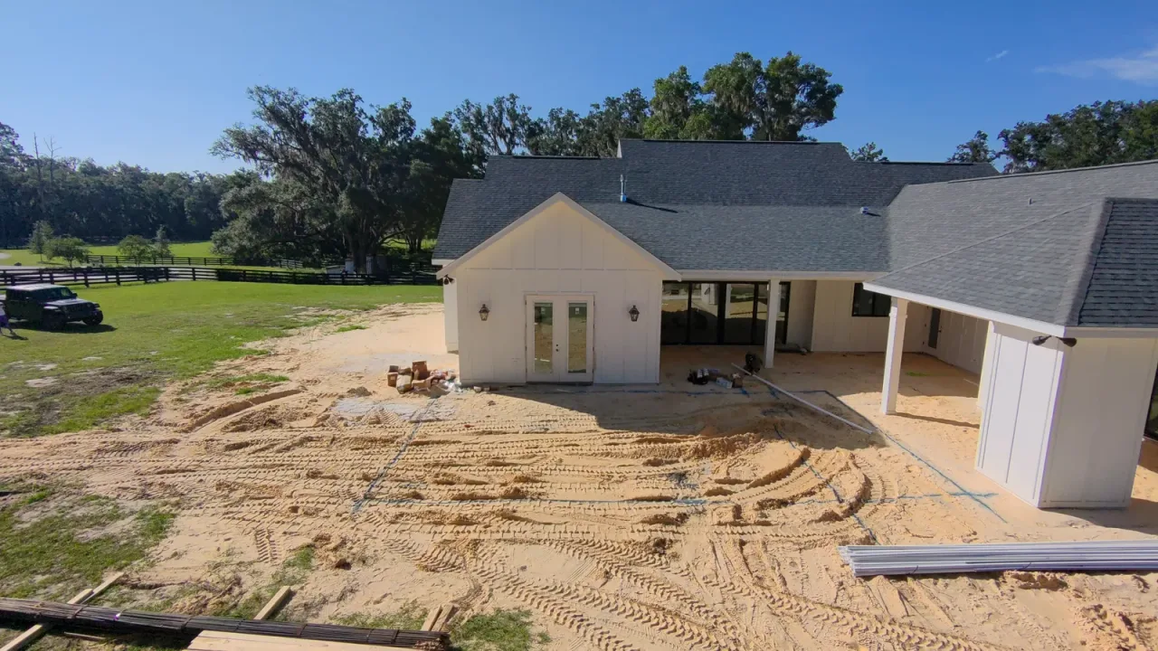 Rear of a white house with blue pool layout lines painted on a sandy, tire-tracked yard, and large trees and pasture behind.